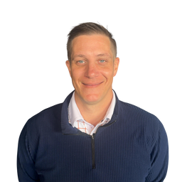 A man smiling, wearing a navy blue textured quarter-zip pullover sweater over a white collared shirt, shot against a plain white background.