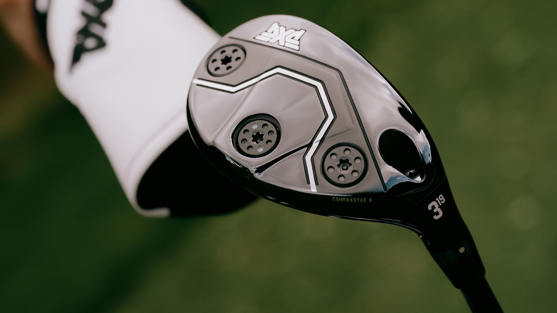 Close-up of a golf club head with visible hexagonal cavity back vents and a white sleeve with black lettering, photographed against a blurred green outdoor background.