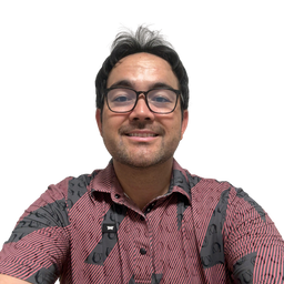 A man with glasses and a short beard smiling, wearing a short-sleeve button-up shirt with a red and black patterned batik design, set against a plain white background.