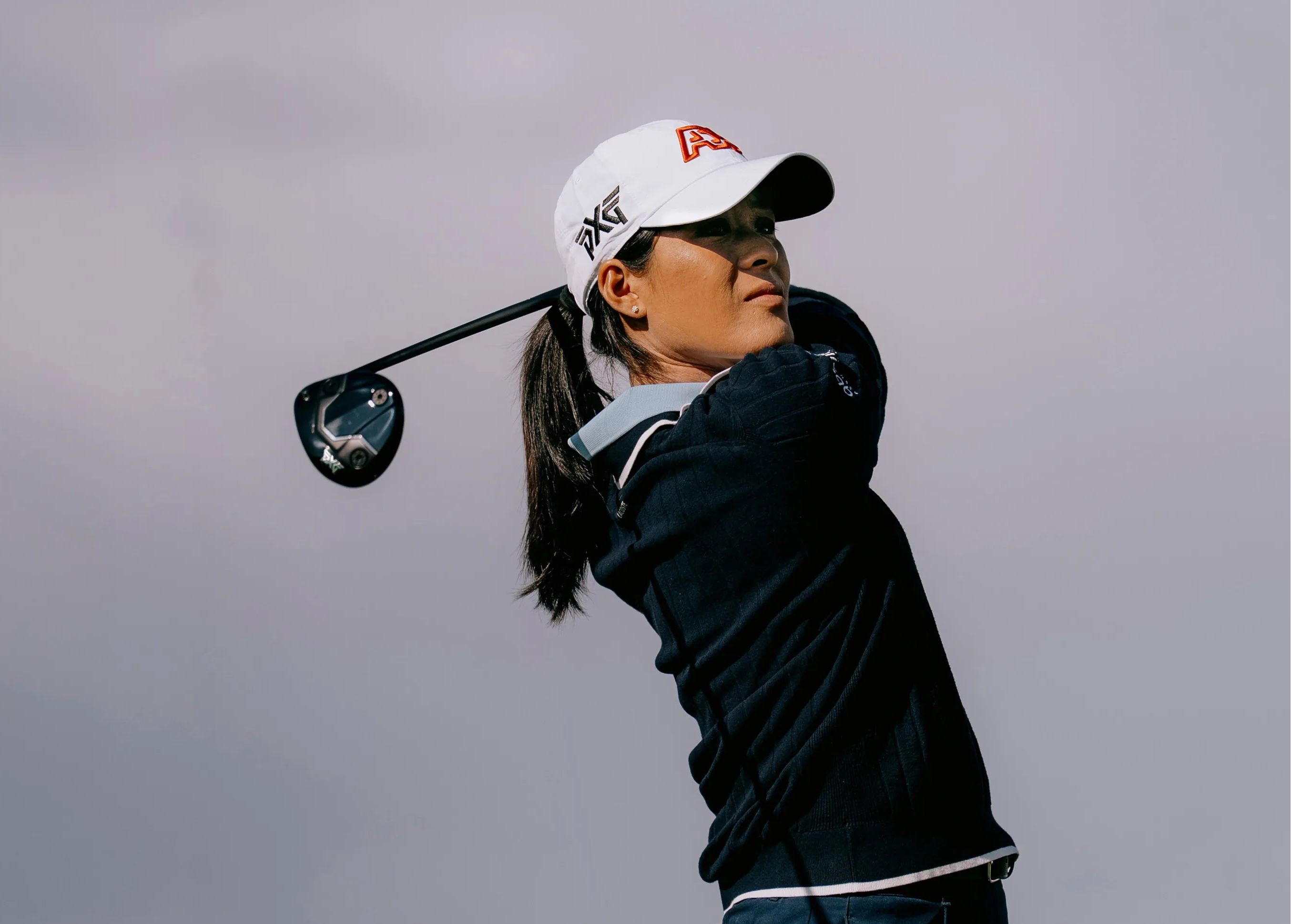 A female golfer in a navy blue outfit and white cap with a red logo is captured mid-swing against a cloudy gray sky.