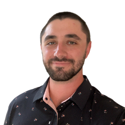 A man with short dark hair and a beard wearing a black patterned button-down shirt over a light pink undershirt, shown against a plain white background.
