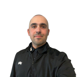 A man with a shaved head wearing a black zip-up jacket over a black collared shirt, photographed against a plain white background.