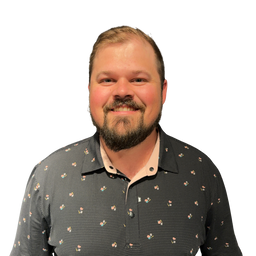 A smiling man with a beard and short hair wearing a dark gray patterned button-down shirt, photographed against a plain white background.