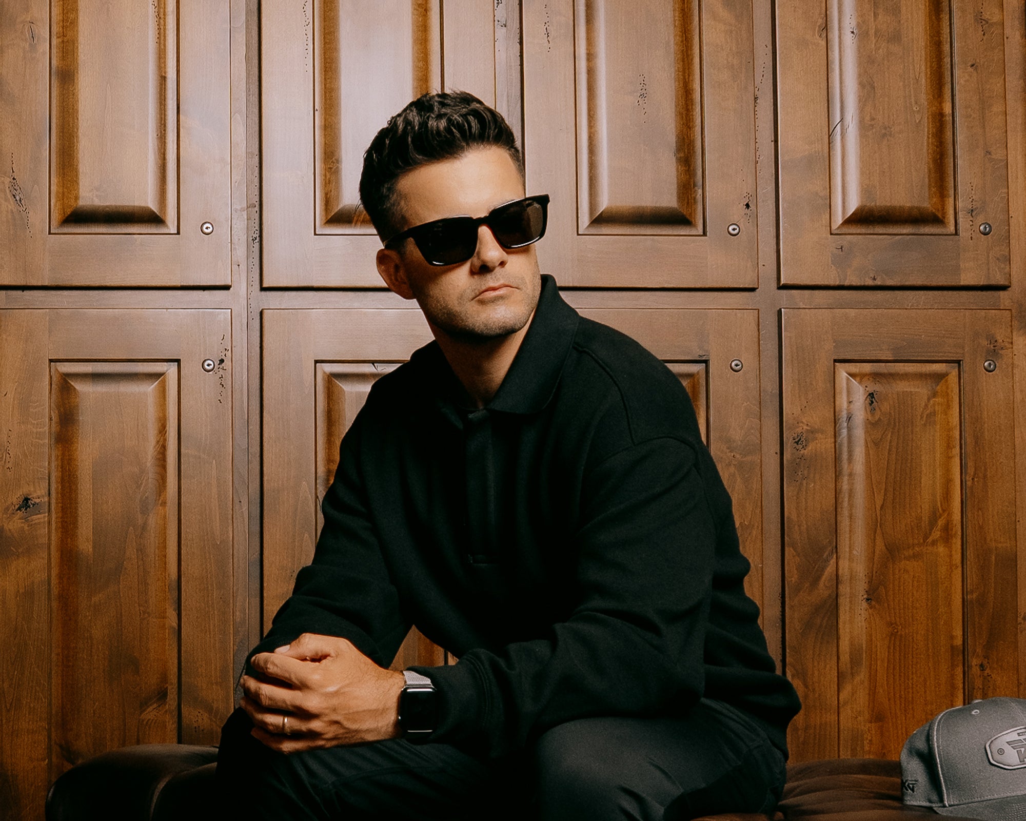 Man wearing sunglasses and a black outfit sitting against a wooden paneled wall.