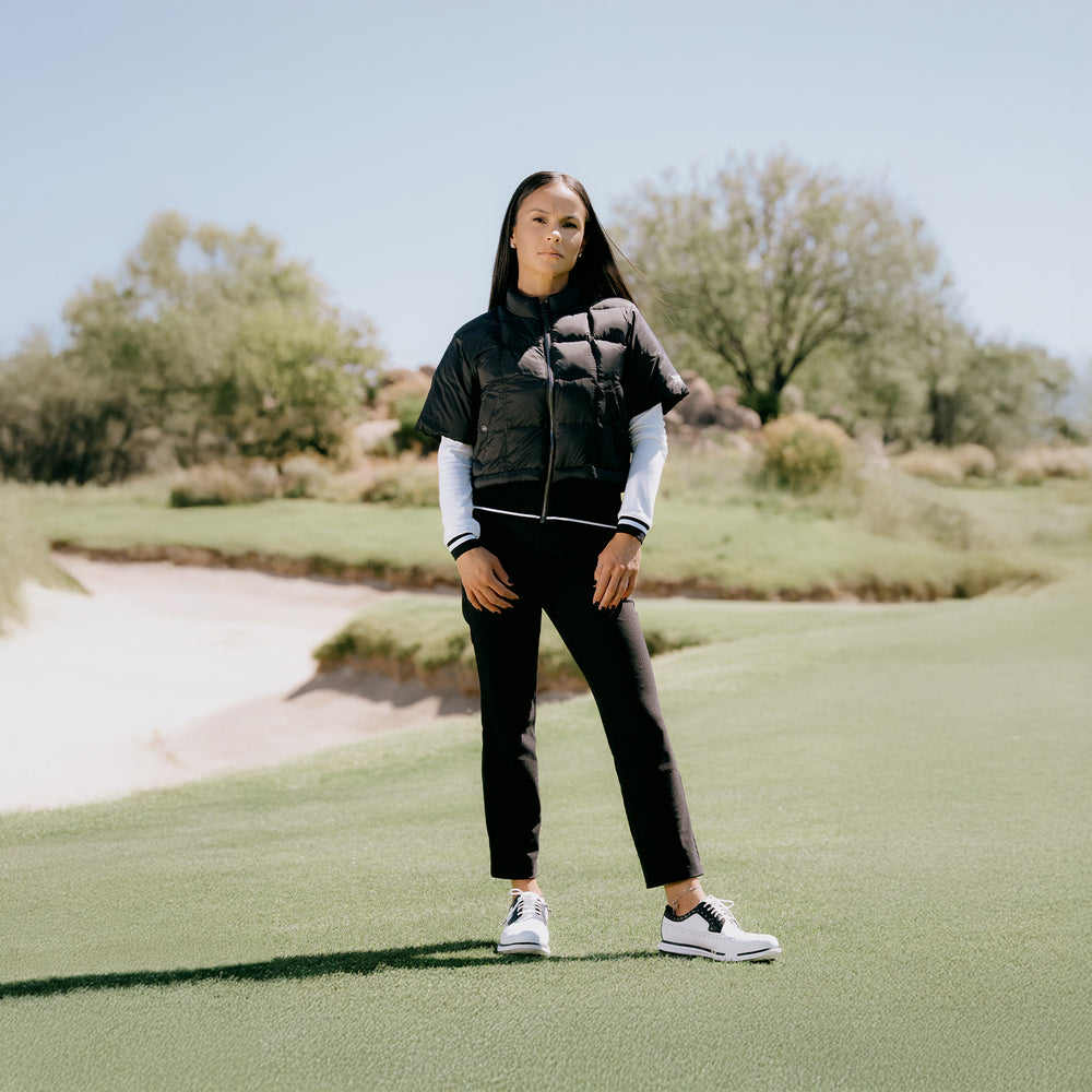 Person standing on a golf course wearing a black and gray jacket, white shirt, black pants, and white shoes.