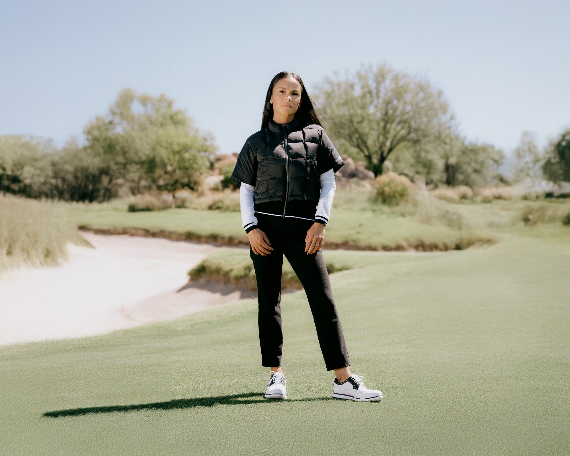 Person standing on a golf course wearing a black and gray jacket, white shirt, black pants, and white shoes.