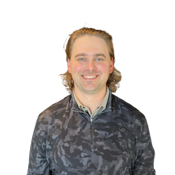 A smiling man with light brown hair wearing a dark gray and black camouflage quarter-zip pullover over a light beige collared shirt, set against a plain white background.