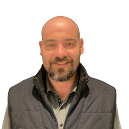 A smiling bald man with a beard wearing a dark gray quilted vest over a light gray button-down shirt, isolated on a white background.