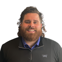 A smiling man with a full beard and curly brown hair wearing a black zip-up athletic jacket over a blue collared shirt, photographed against a plain white background. A small white logo is visible on the left chest of the jacket.