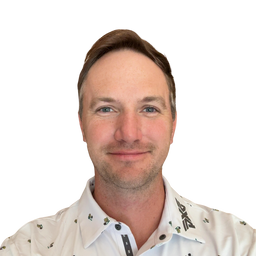 A man with light blue eyes and brown hair wearing a white button-down shirt with a small green and black pattern, set against a plain white background.
