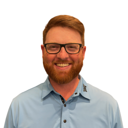 A smiling man with a reddish-brown beard and glasses wearing a light blue button-down shirt, shown against a plain white background.