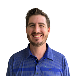 A smiling man with short brown hair and a beard wearing a blue patterned button-down shirt, photographed against a plain white background.