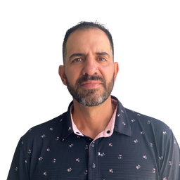 A man with a beard and short dark hair wearing a dark navy patterned button-down shirt over a light pink collared undershirt, photographed against a plain white background.