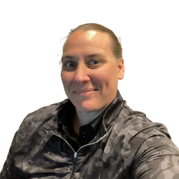 A close-up portrait of a woman with her hair pulled back, wearing a dark gray and black camouflage zip-up athletic jacket over a black shirt, set against a solid white background.