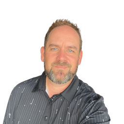 A man with short brown hair and a beard wearing a dark patterned button-up shirt, photographed against a plain white background.