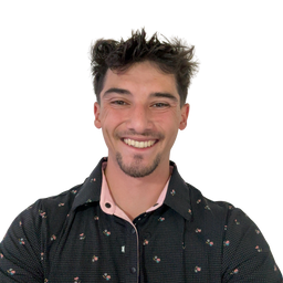 A smiling young man with dark hair and a short beard, wearing a black patterned button-up shirt over a light pink undershirt, photographed against a plain white background.