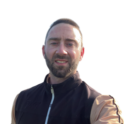 A man wearing a black zip-up vest over a tan long-sleeve shirt, photographed outdoors against a bright white sky.