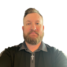 A man with a beard and styled hair wearing a black button-up shirt with a gray collar and a silver cross necklace, photographed against a plain white background.