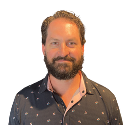 A man with a beard wearing a dark patterned button-down shirt with a light pink inner collar, photographed against a plain white background.
