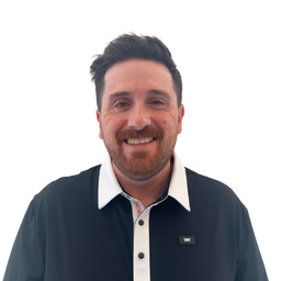 A smiling man with short dark hair and a beard wearing a black polo shirt with a white collar, photographed against a plain white background.