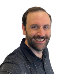 A smiling man with a beard wearing a dark patterned button-up shirt, photographed against a plain white background.