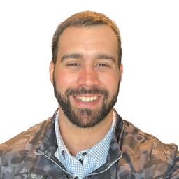 A smiling man with a beard wearing a camouflage-patterned zip-up jacket over a light blue patterned collared shirt, set against a plain white background.