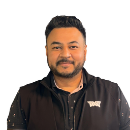 A man with a beard and styled hair wearing a black zip-up vest over a dark patterned shirt, featuring a white stylized logo on the left chest, set against a plain white background.