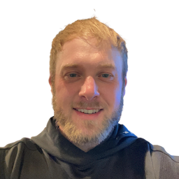 A close-up portrait of a smiling man with blond hair and a beard, wearing a dark gray hoodie against a plain white background.