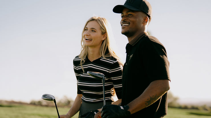 A smiling blonde woman in a black and white striped polo shirt stands next to a man in a black polo shirt and cap on a golf course, holding golf clubs.
