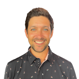 A smiling man with short brown hair and light blue eyes wearing a dark patterned button-down shirt, photographed against a plain white background.