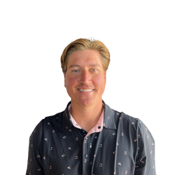 A smiling man with blonde hair wearing a dark patterned button-up shirt over a light pink collared shirt, photographed against a plain white background.