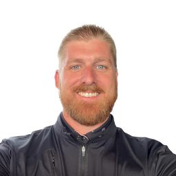 A smiling man with short blond hair and a reddish beard, wearing a dark navy zip-up jacket over a patterned collared shirt, photographed against a plain white background.