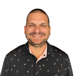A smiling man with short dark hair and a trimmed beard, wearing a black short-sleeve button-up shirt with a small multicolor floral pattern over a light pink inner collar, photographed against a plain white background.