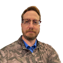 A man with a beard and glasses wearing a camouflage zip-up jacket over a blue collared shirt, photographed against a plain white background.