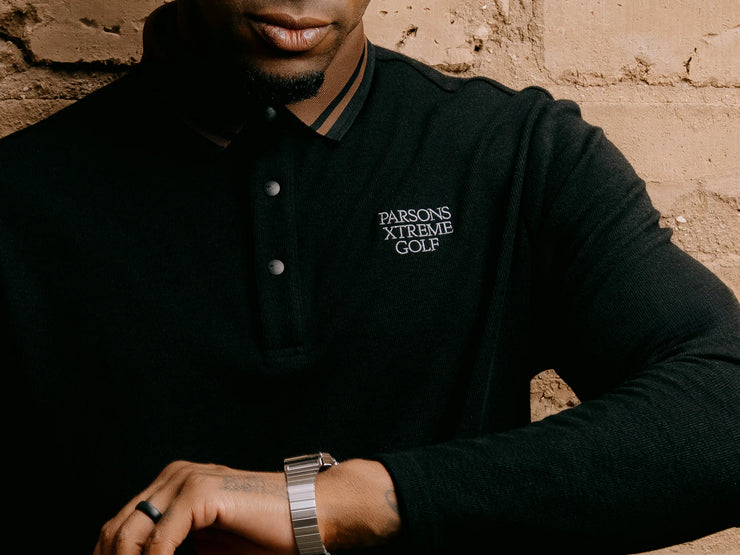 A close-up of a man wearing a black long-sleeve polo shirt with 'PARSONS XTREME GOLF' embroidered in white on the left chest, posed against a textured tan brick wall.
