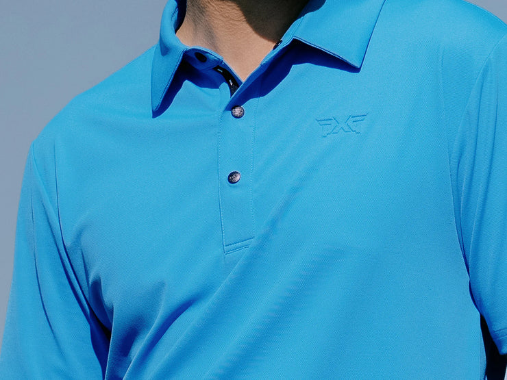 A close-up of a person wearing a bright blue long-sleeve polo shirt with a subtle tonal logo on the left chest, set against a pale blue-gray background.