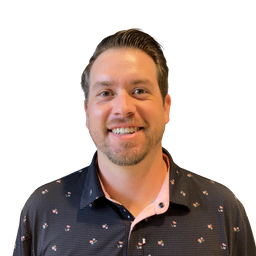 A smiling man with short brown hair and light facial stubble, wearing a dark patterned button-up shirt over a light pink undershirt, photographed against a plain white background.