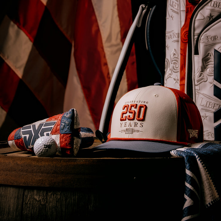 Cap with 'Celebrating 250 Years' text, golf club, and golf ball on a wooden surface with American flag in the background