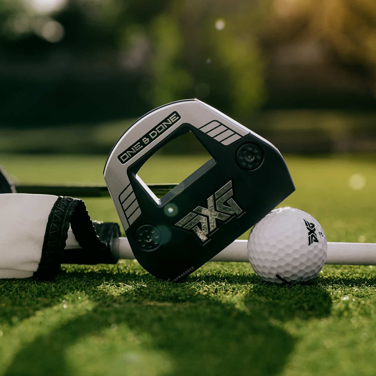 A close-up of a PXG One 6 Dune golf club head resting on a golf ball on a green grass surface, with a blurred outdoor golf course background.