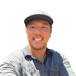 A smiling man wearing a gray and navy long-sleeve button-up shirt and a gray cap, shown in a close-up selfie-style portrait against a bright white background.