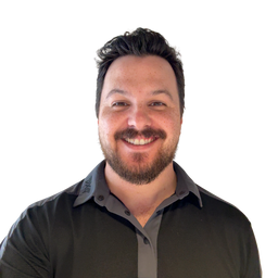 A smiling man with dark hair and a beard wearing a dark gray button-up shirt, photographed against a plain white background.
