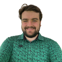 A smiling man with brown hair and a beard wearing a green patterned button-down shirt, photographed against a plain white background.