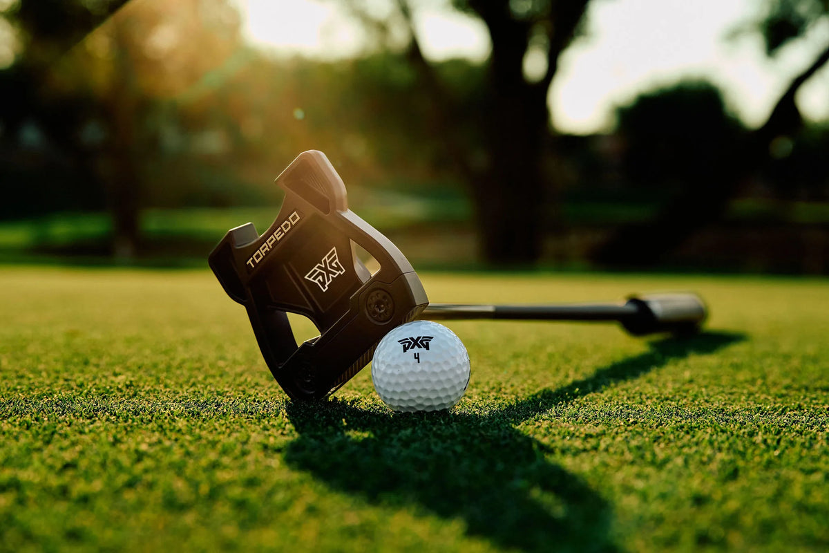 A Torpedo golf putter head resting on a golf green next to a white golf ball, with a blurred outdoor golf course background and warm sunlight.