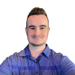 A man wearing a blue and purple patterned button-down shirt, photographed in a selfie-style close-up against a plain white background.