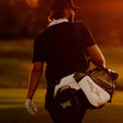 A golfer seen from behind walking on a golf course at sunset, carrying a white and black golf bag over his shoulder.