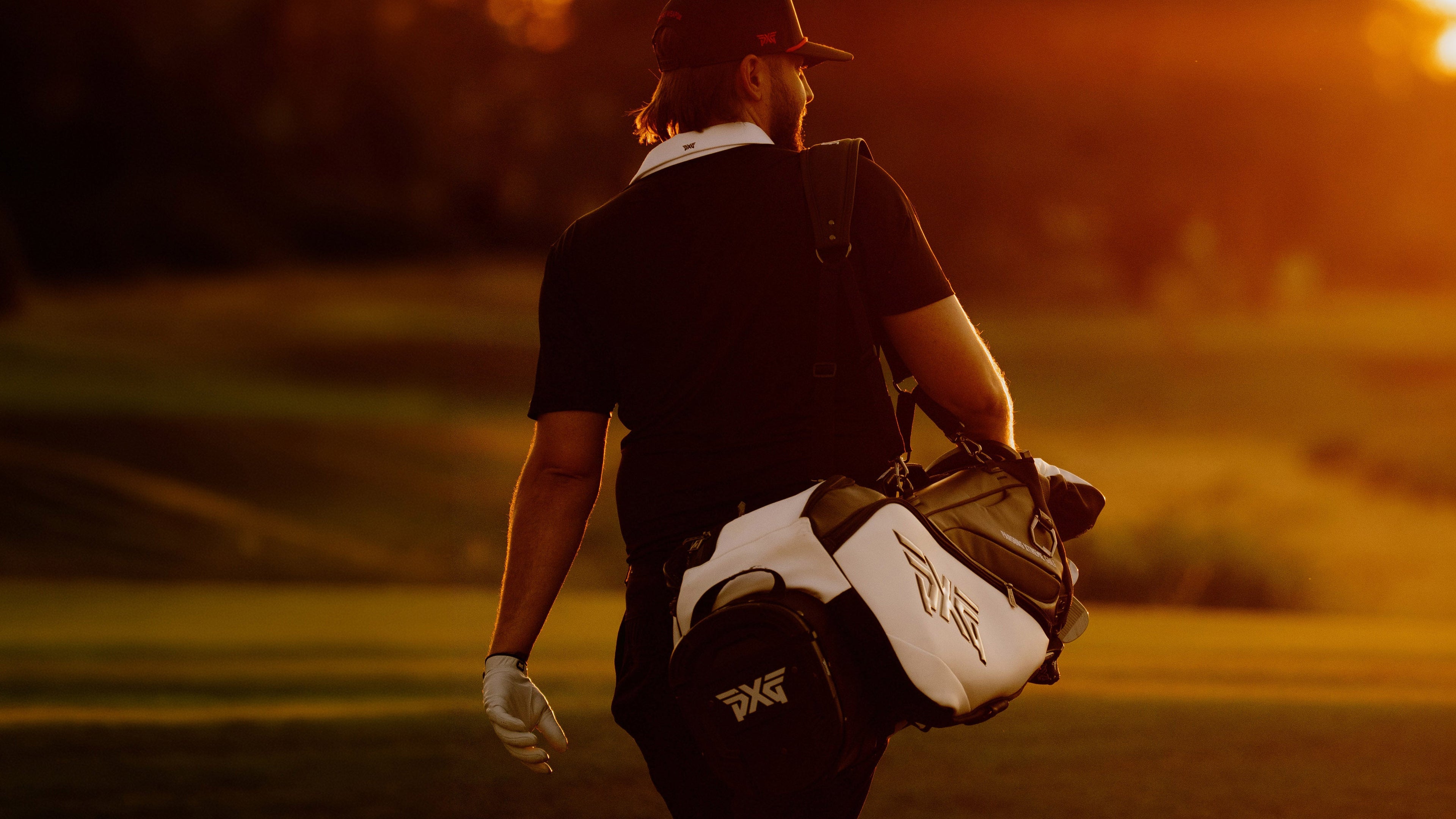 A golfer seen from behind walking on a golf course at sunset, carrying a white and black golf bag over his shoulder.