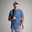 Man wearing a blue polo shirt and gray pants, holding a black backpack against a gray background